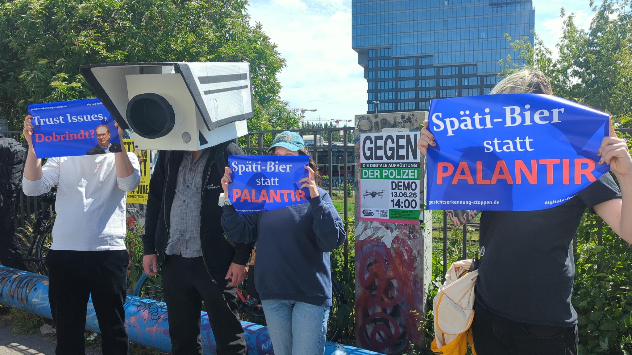 Demonstrierende halten Protestschilder in die Kamera. Eine Kamera schaut zurück.