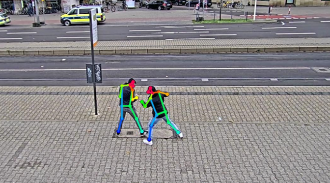 Foto einer Straßenszene schräg von oben aufgenommen, Tag. Im Vordergrund zwei Personen in Interaktion, markiert durch bunte Streifen. Im Hintergrund quer verlaufende Straßen. Quelle Polizei Mannheim.